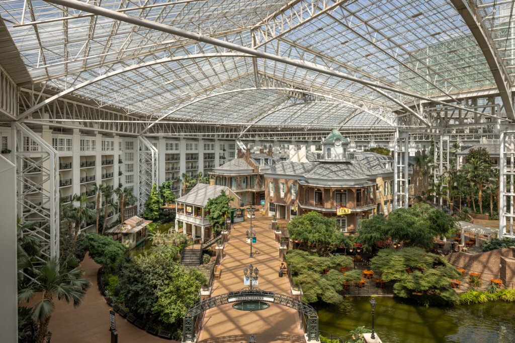 Gaylord Opryland - Expansive glass-roofed atrium featuring replica historic buildings, lush tropical vegetation, a pedestrian plaza with bridge over water, and tall steel framework overhead