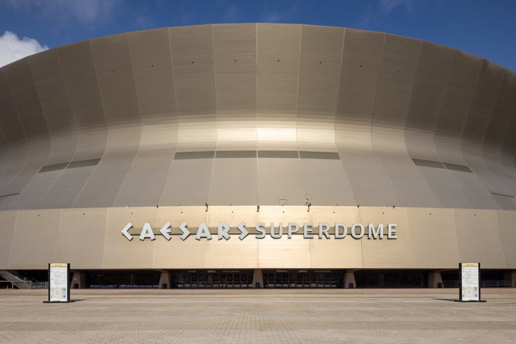 New Orleans Superdome - Large curved beige dome structure with vertical ribbed panels and illuminated signage reading Caesars Superdome, wide plaza in foreground under clear blue sky