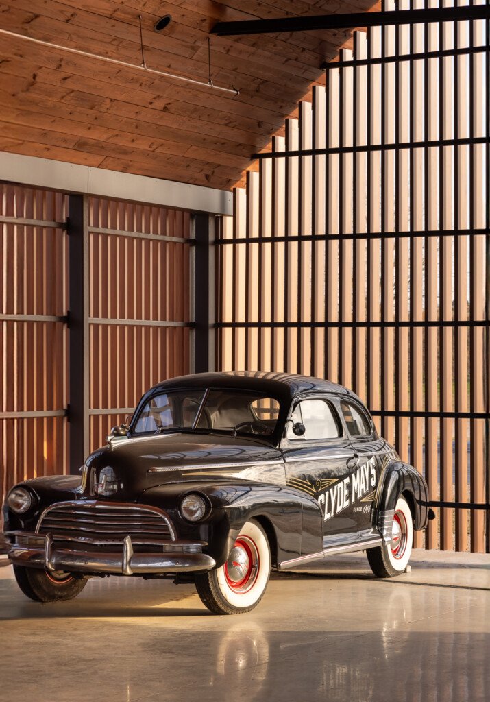 Clyde May's Distillery - A black 1940s coupe with red-wall tires sits in a concrete garage with exposed wood ceiling and illuminated vertical slat walls.