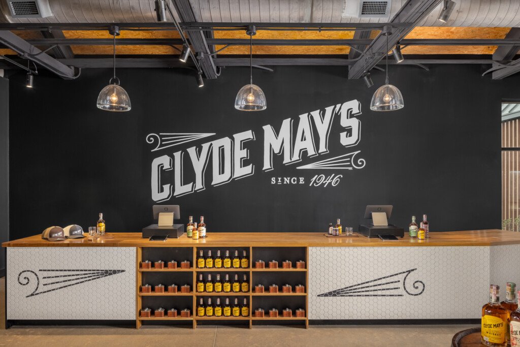 Clyde May's Distillery - Modern bar interior with wooden beam ceiling, pendant lighting, black accent wall with Clyde May's logo, and white tile bar front displaying whiskey bottles.