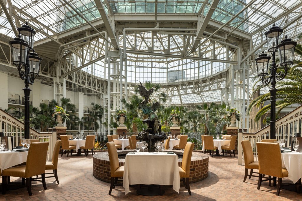 Elegant dining room with round white-clothed tables, tan upholstered chairs, ornamental street lamps, and abundant green plants beneath an expansive glass-paneled roof structure