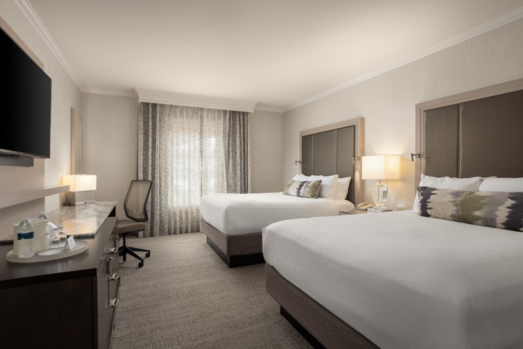 Spacious hotel room featuring two queen beds with white linens, desk with office chair, wall-mounted TV, and large windows with layered curtains