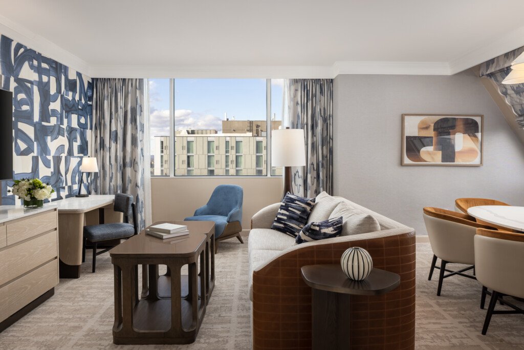 Loews Vanderbilt - Bright hotel suite with curved sofa, blue patterned curtains, wood furniture, and floor-to-ceiling windows overlooking a city street
