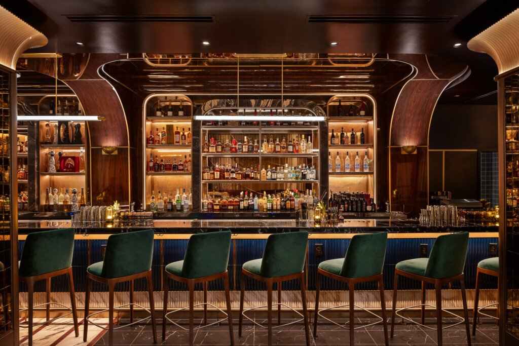 Philippe Chow - Nashville - Dark wood bar with illuminated shelving displaying spirits, green upholstered bar stools, neon accents, and decorative wooden architectural details.