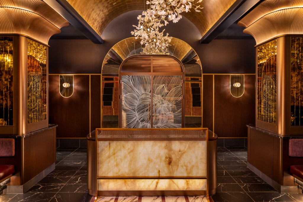 Philippe Chow - Nashville - A symmetrical restaurant lobby features a marble reception desk with brass details, ornate ceiling with curved beams and a floral chandelier, rich brown walls with gold accents, and decorative sconces flanking an artistic floor pattern.