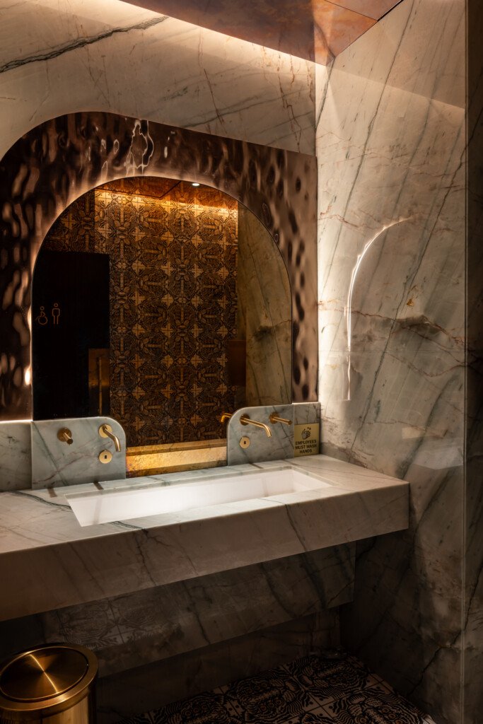 Philippe Chow - Nashville - Marble bathroom with large arched mirror, white sink, brass fixtures, patterned gold wallpaper, and ambient lighting