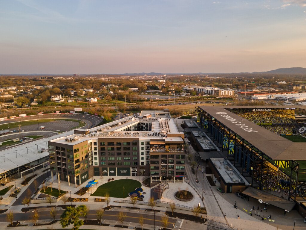 Aerial view of a modern mixed-use building with yellow and white facades, rooftop plaza, surrounding parking lots, sports stadium, and forested residential areas under evening light.