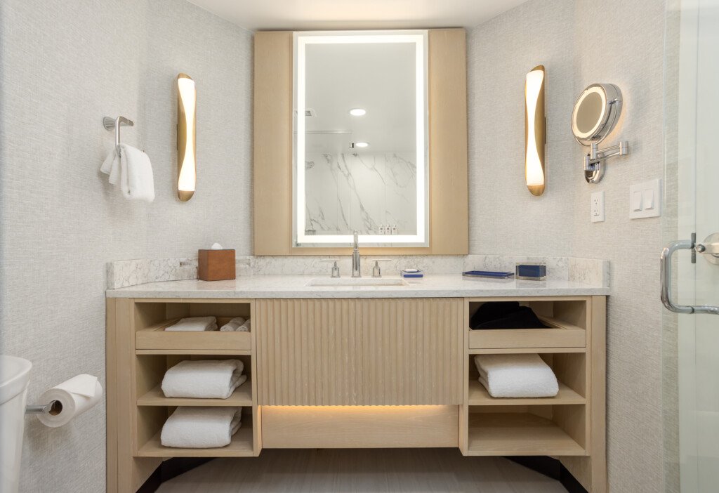 Light wood vanity with open shelving, marble countertop, lighted rectangular mirror, brass sconces, and organized towel storage