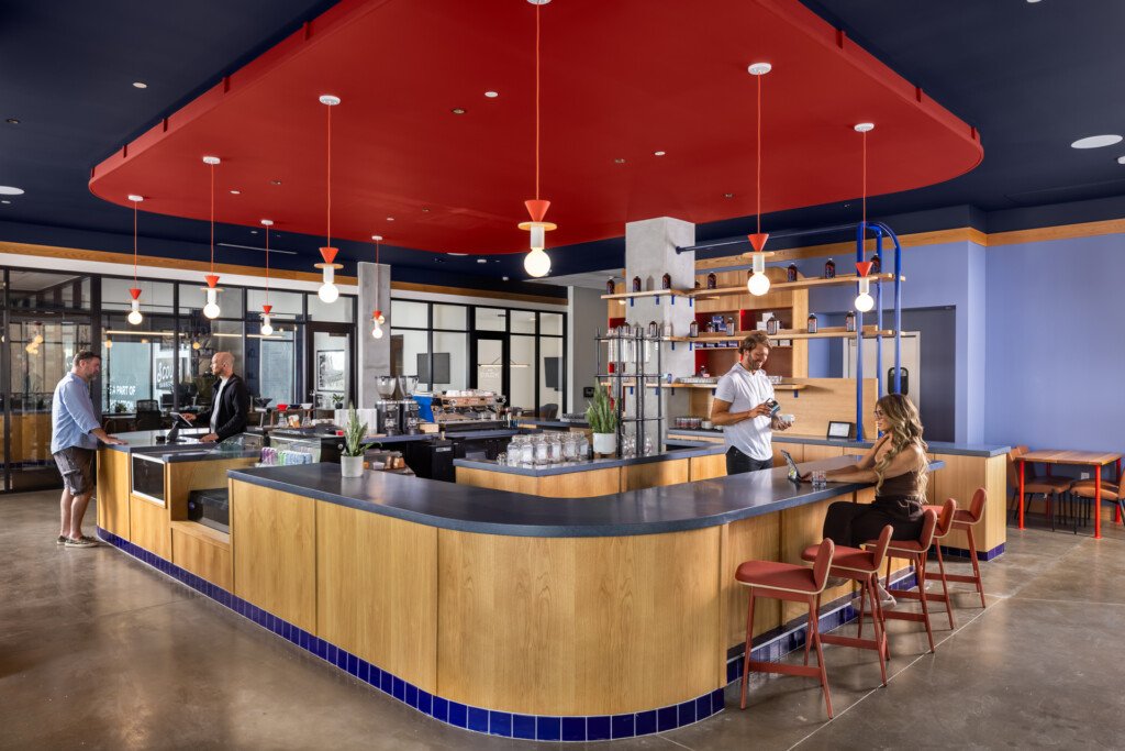 Open-concept kitchen with curved wooden service counter, red suspended ceiling, pendant lighting, and seating area with guests and staff interacting.