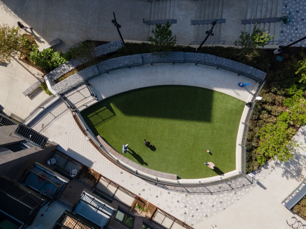 Overhead view of a curved courtyard with lush green lawn, concrete seating areas, metal benches, and people relaxing on the grass surrounded by building walls and landscaping