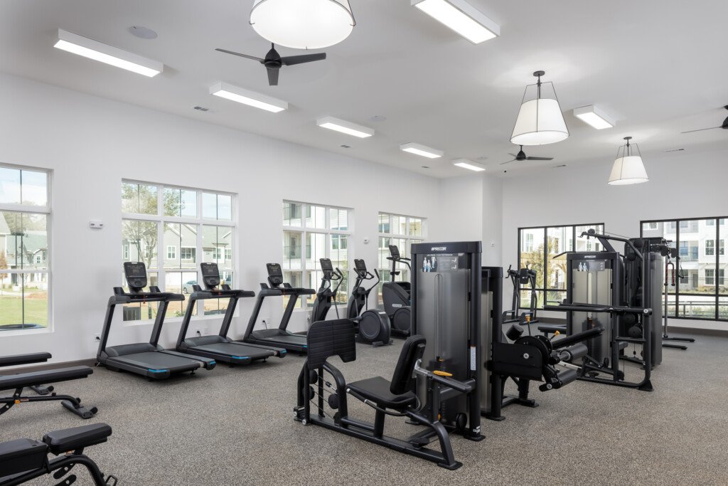 Bright fitness center with rows of treadmills, exercise machines, pendant lighting, and large windows overlooking landscaped grounds
