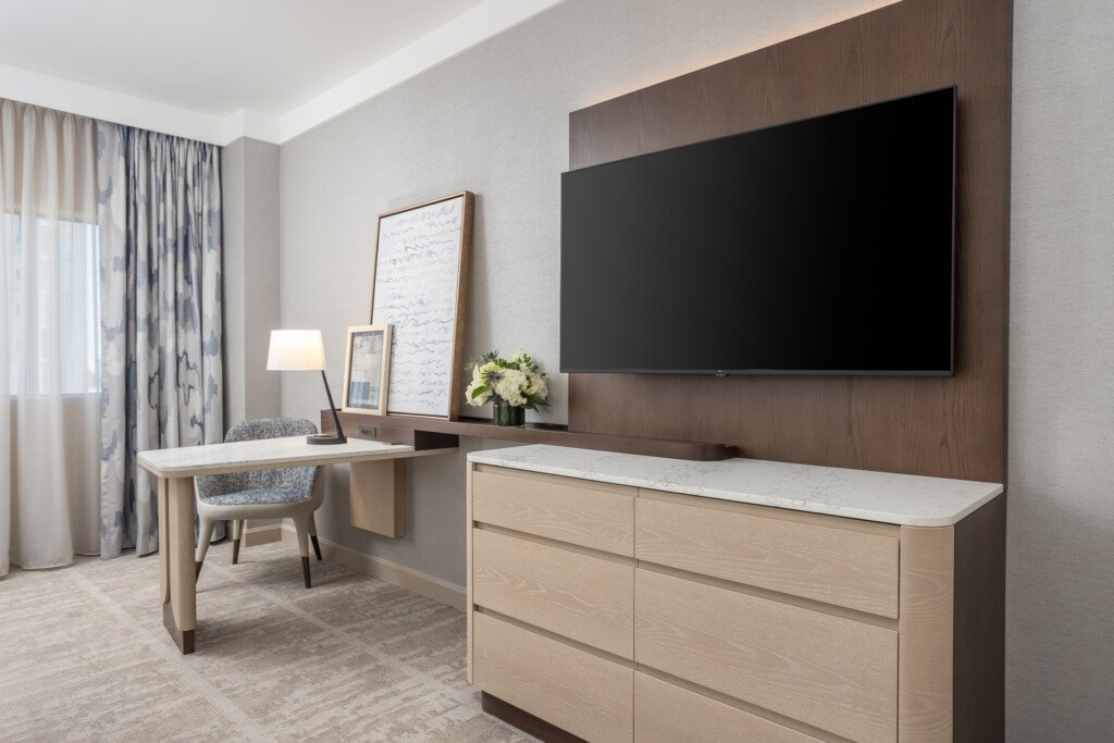 Loews Vanderbilt - Hotel room interior featuring a wall-mounted TV on a dark wood panel, light wood dresser with marble countertop, desk with chair by window, and framed artwork on the wall