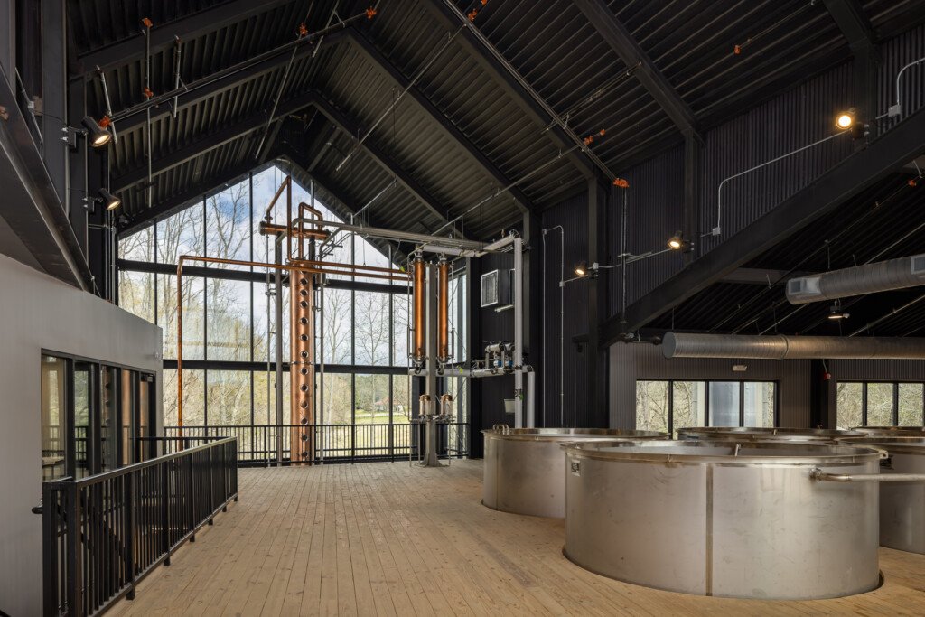 Clyde May's Distillery - Large metal fermentation tanks in a brewery space with black metal railings, dramatic vaulted corrugated ceiling, and expansive windows overlooking bare trees.