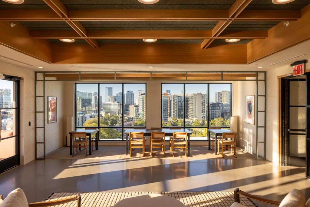Bright commercial lounge with warm wood ceiling beams, floor-to-ceiling windows overlooking city skyline, wooden chairs, polished concrete flooring, and recessed lighting.