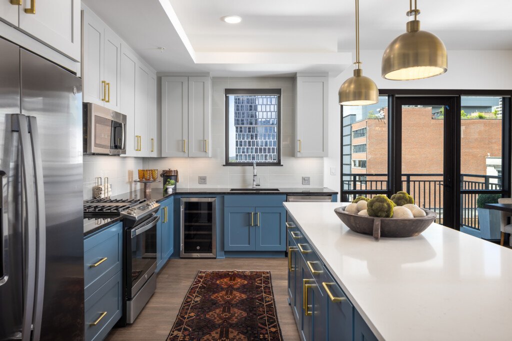 Contemporary kitchen with white upper cabinets, blue lower cabinets, stainless steel appliances, white countertops, brass pendant lights, and black-framed glass doors opening to a balcony.