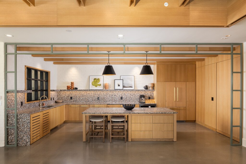 Contemporary kitchen with natural wood cabinetry, terrazzo backsplash, green-framed glass doors, central island with seating, and pendant lighting overhead.