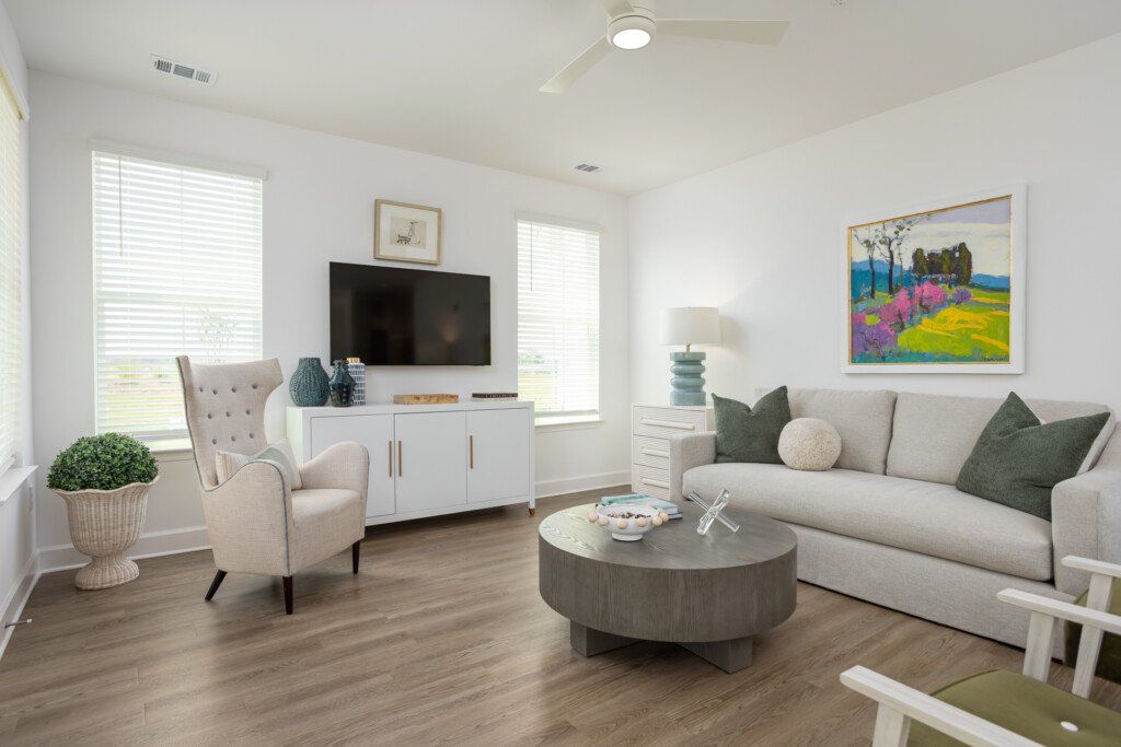 Bright living room with cream sofa, beige accent chair, oval wood coffee table, wall-mounted TV, and colorful landscape artwork on walls