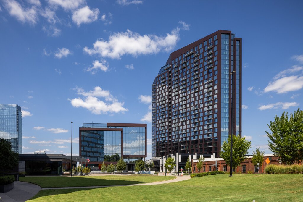 Tall residential building with brick and glass facade, contemporary mid-rise structures, and green lawn plaza under blue sky