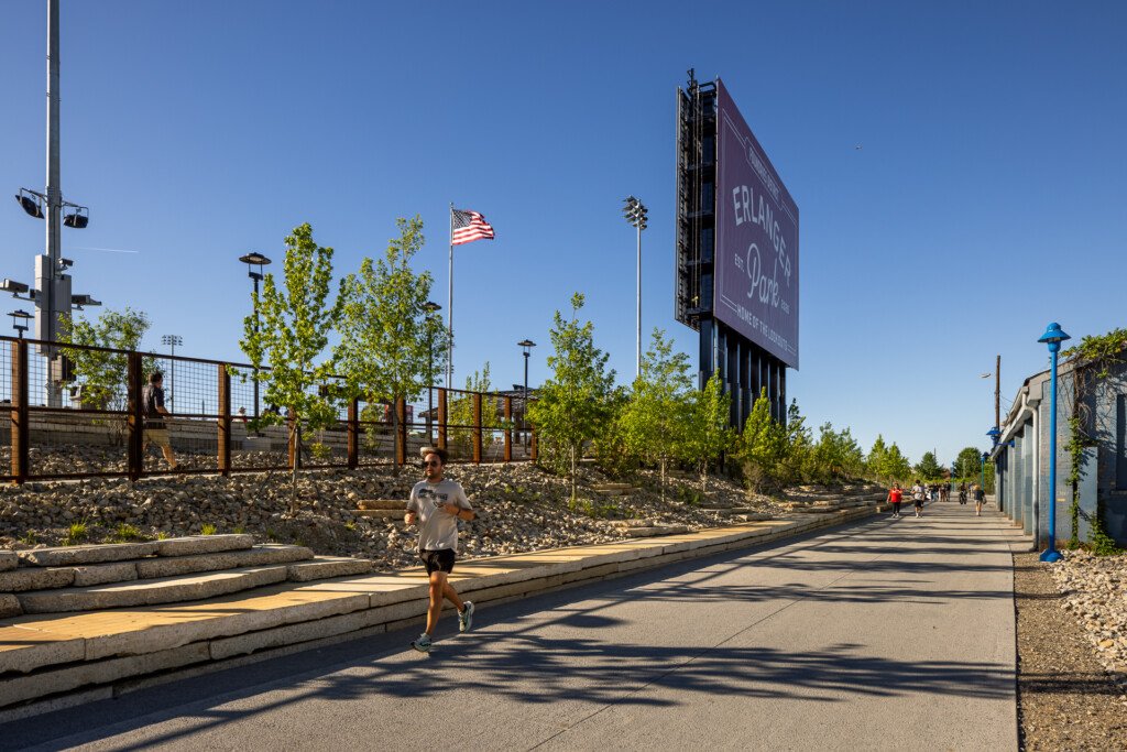 Runner jogging on a paved path through a contemporary public park with wooden log seating, landscaped hillside, American flag, and large digital display board under clear blue sky