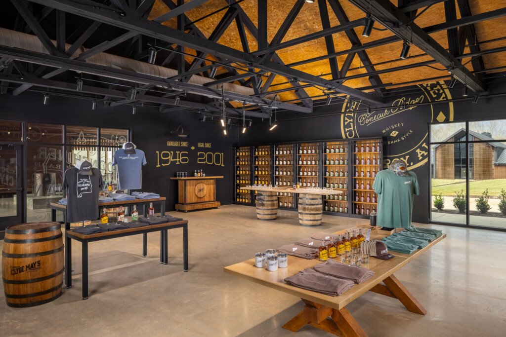 Clyde May's Distillery - Industrial tasting room with black steel beam ceiling, golden skylights, bourbon barrel displays, retail shelving, and merchandise tables showcasing spirits and branded apparel.
